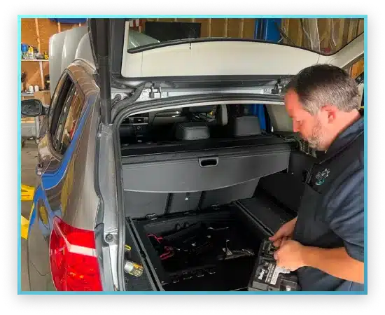 A mechanic works on a car in a workshop, accessing the trunk area with various tools and equipment visible around.